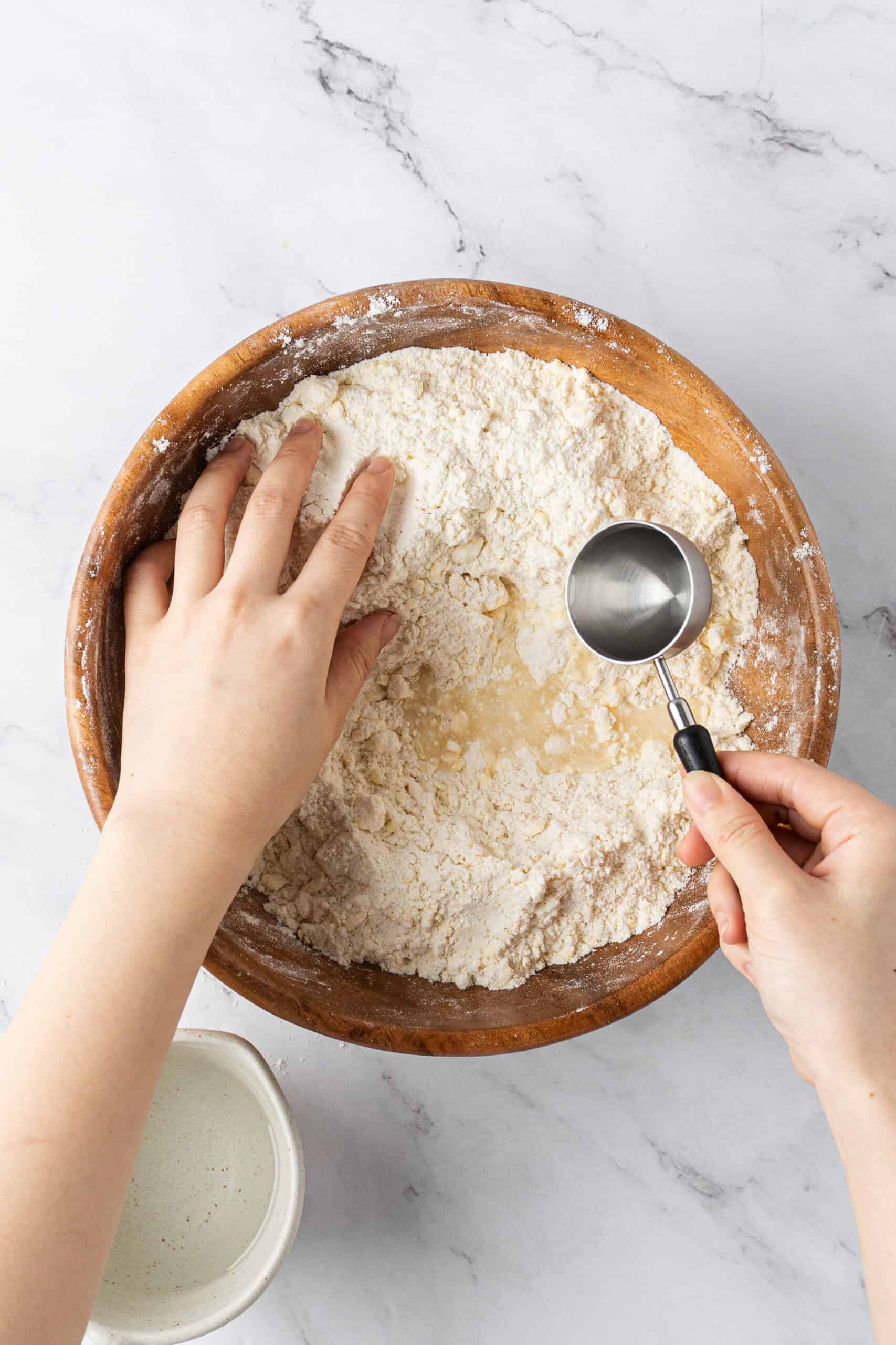 Adding water to a crumbly flour and butter mixture in a wooden bowl to make pie crust dough.