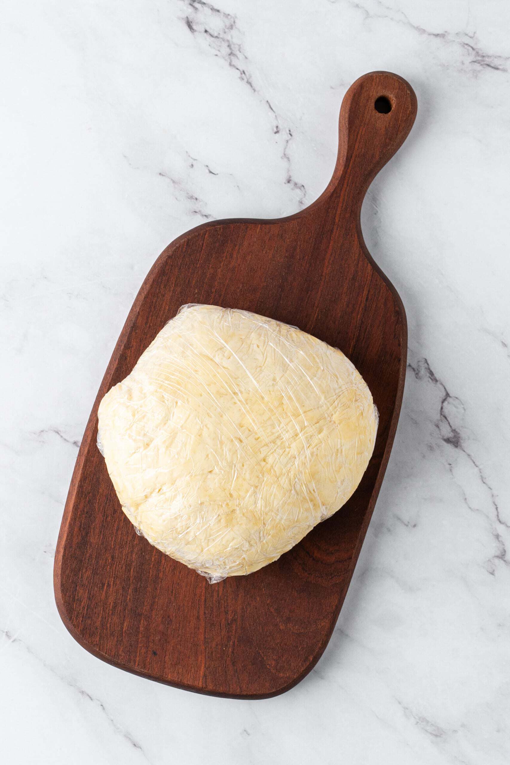 Homemade pie crust dough wrapped in plastic wrap and resting on a dark wooden cutting board.