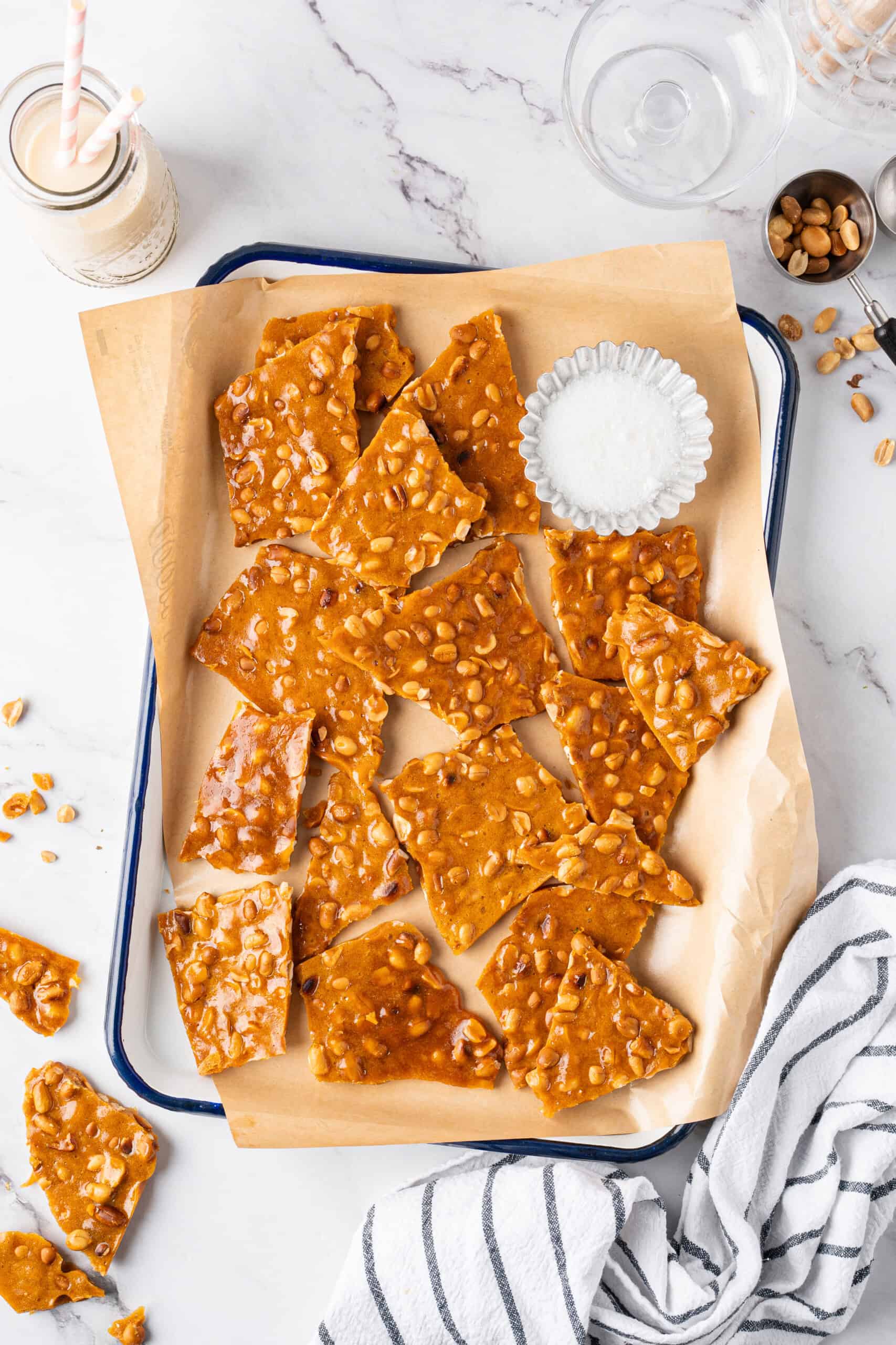 Overhead view of homemade peanut brittle broken into golden pieces on a parchment-lined tray with a bowl of sugar and scattered peanuts.