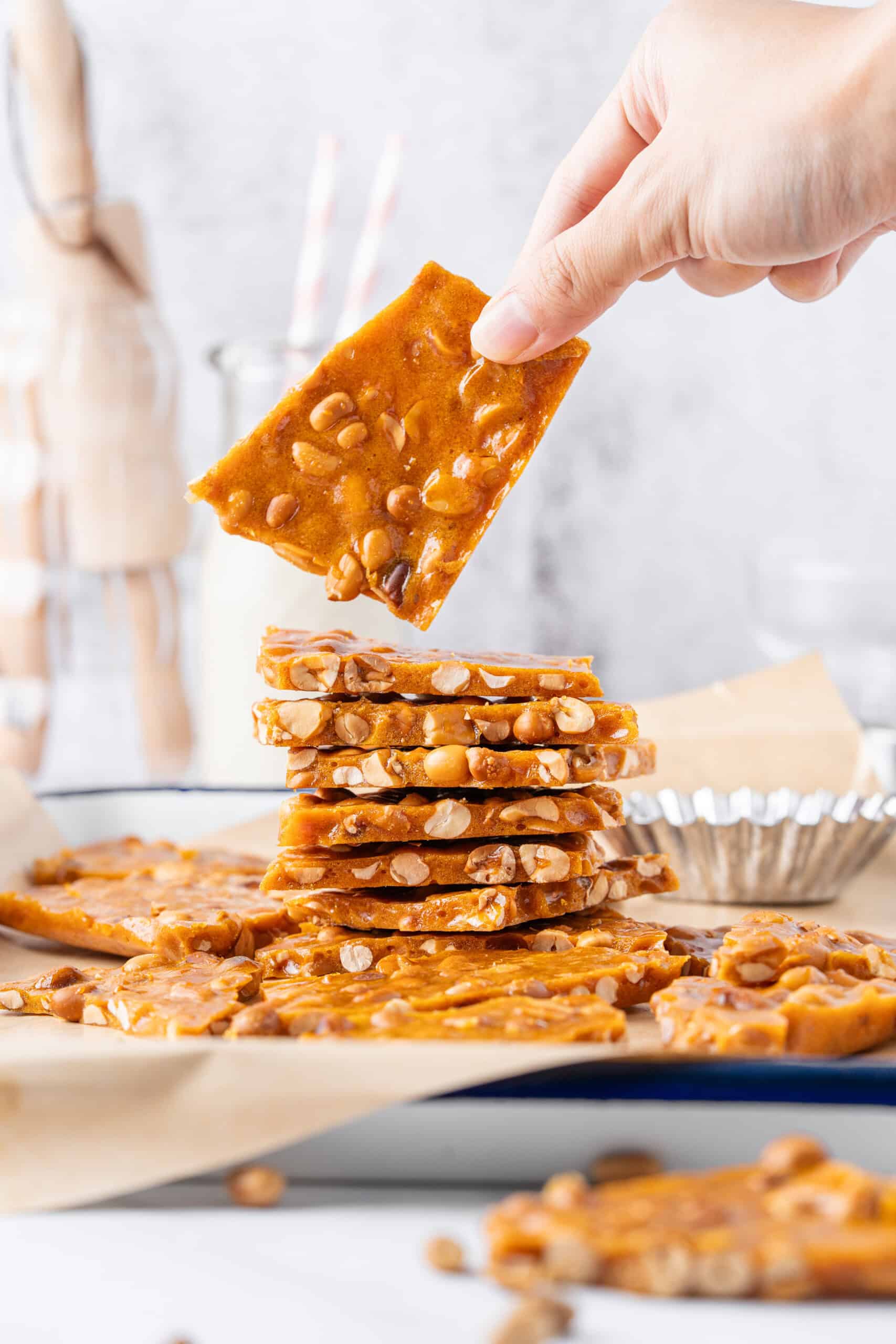 Hand holding a piece of golden homemade peanut brittle above a stack of thin, peanut-filled candy pieces.