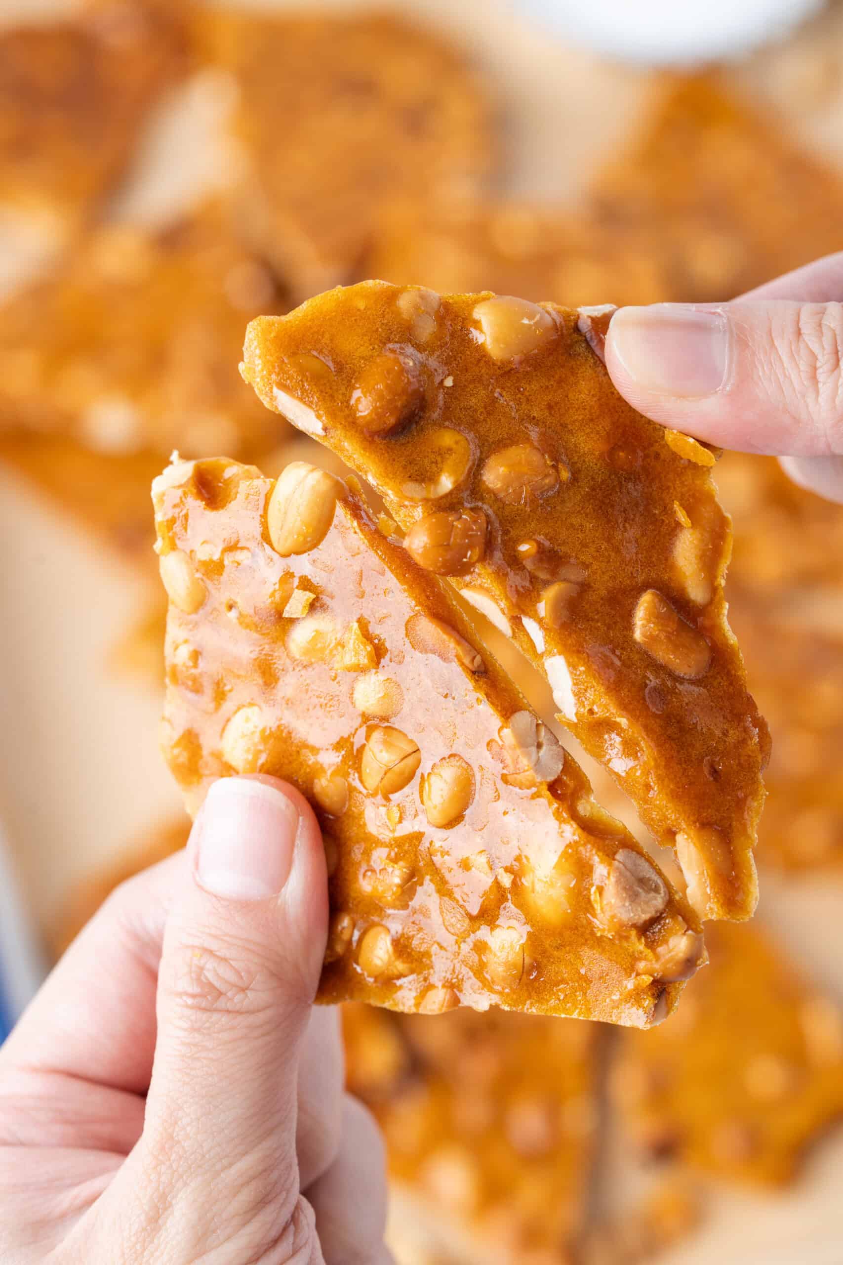 Close-up of hands breaking apart two pieces of golden homemade peanut brittle filled with peanuts.