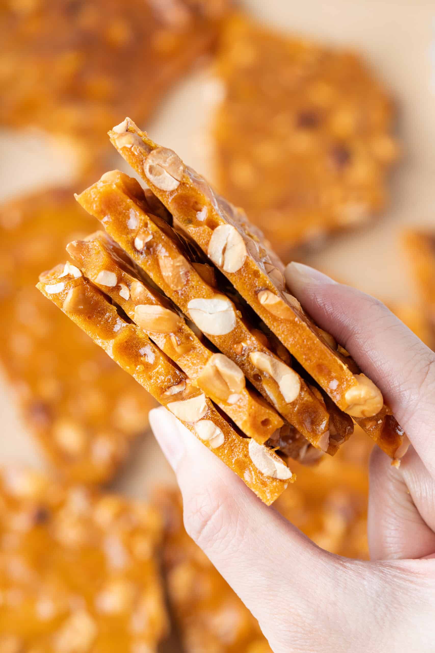 Hand holding several thin pieces of homemade peanut brittle, showing the golden caramel and peanuts throughout.