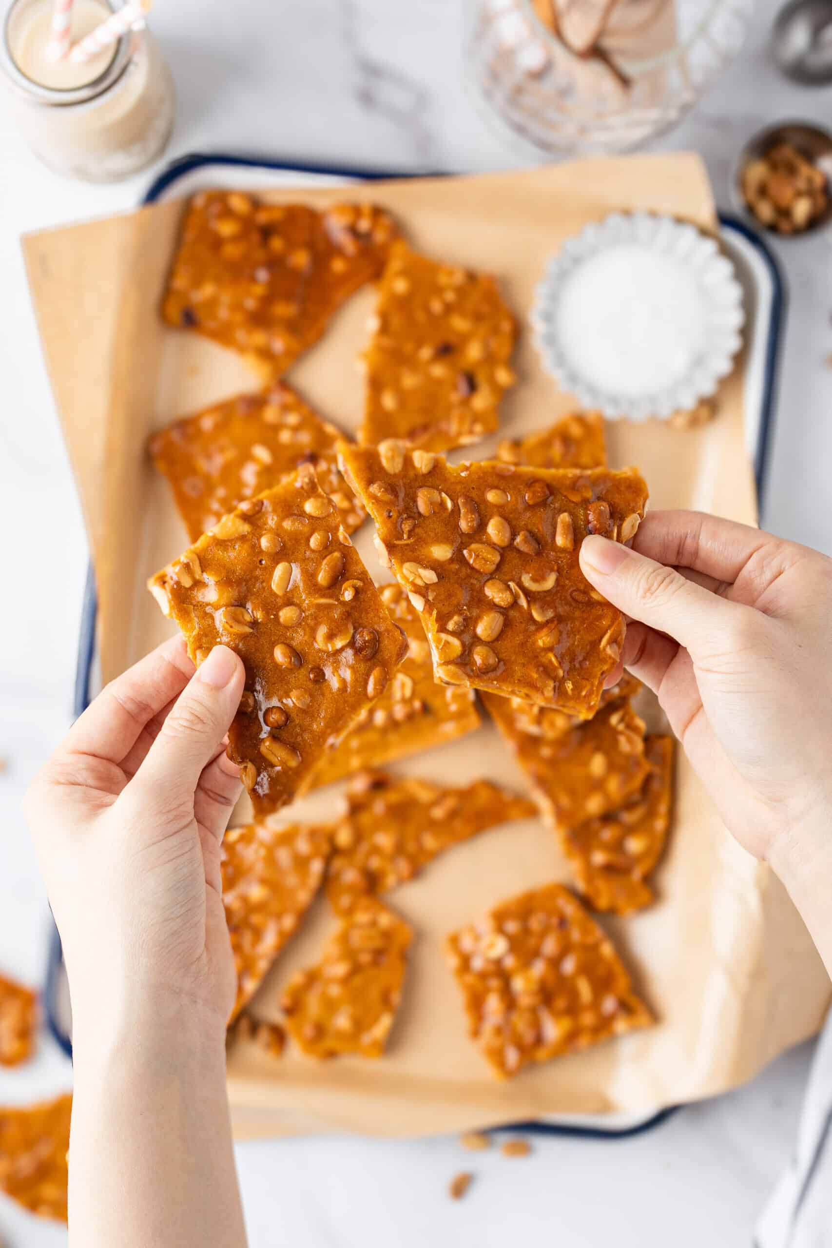 Hands holding two pieces of homemade peanut brittle over a tray of golden, peanut-filled candy.