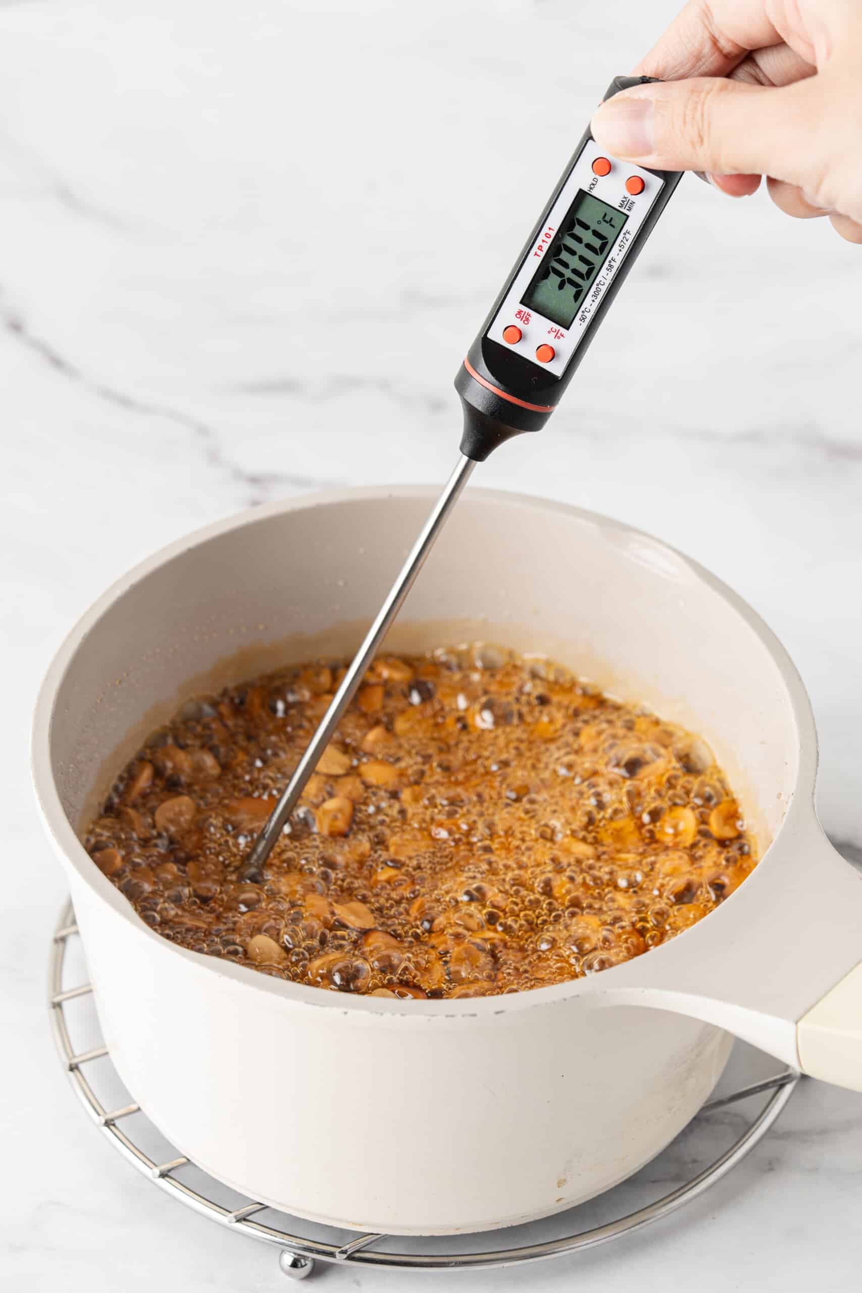 Digital candy thermometer checking the temperature of bubbling golden sugar syrup and peanuts in a saucepan.