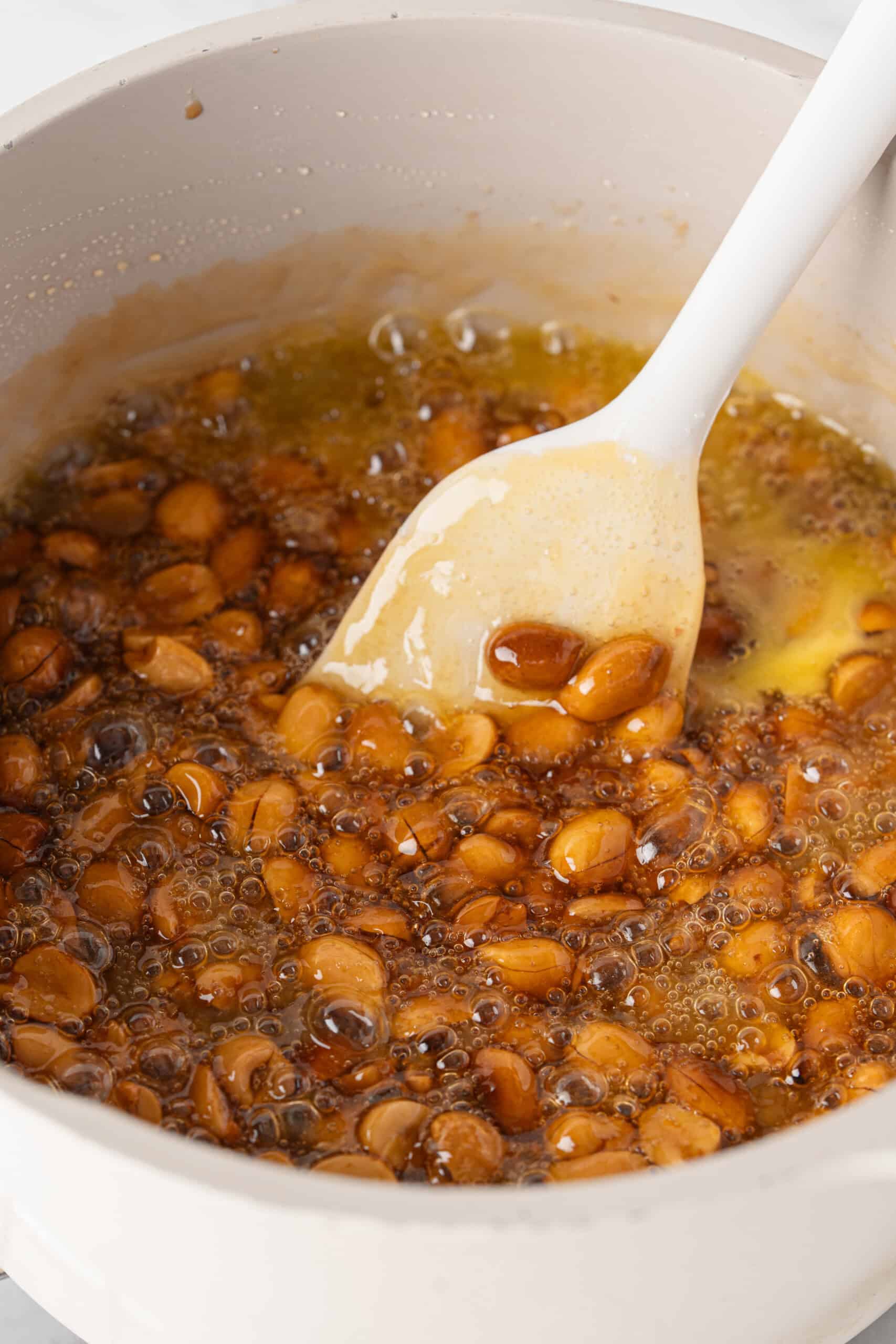 Peanuts coated in bubbling golden caramel mixture being stirred with a white spatula.