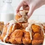 A hand pulling a cinnamon sugar-coated piece of pumpkin monkey bread topped with thick vanilla glaze from the Bundt-shaped loaf.