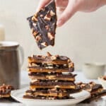 Homemade Toffee with layers of buttery caramel, rich chocolate, and chopped pecans, with one piece held above a stack on a plate.