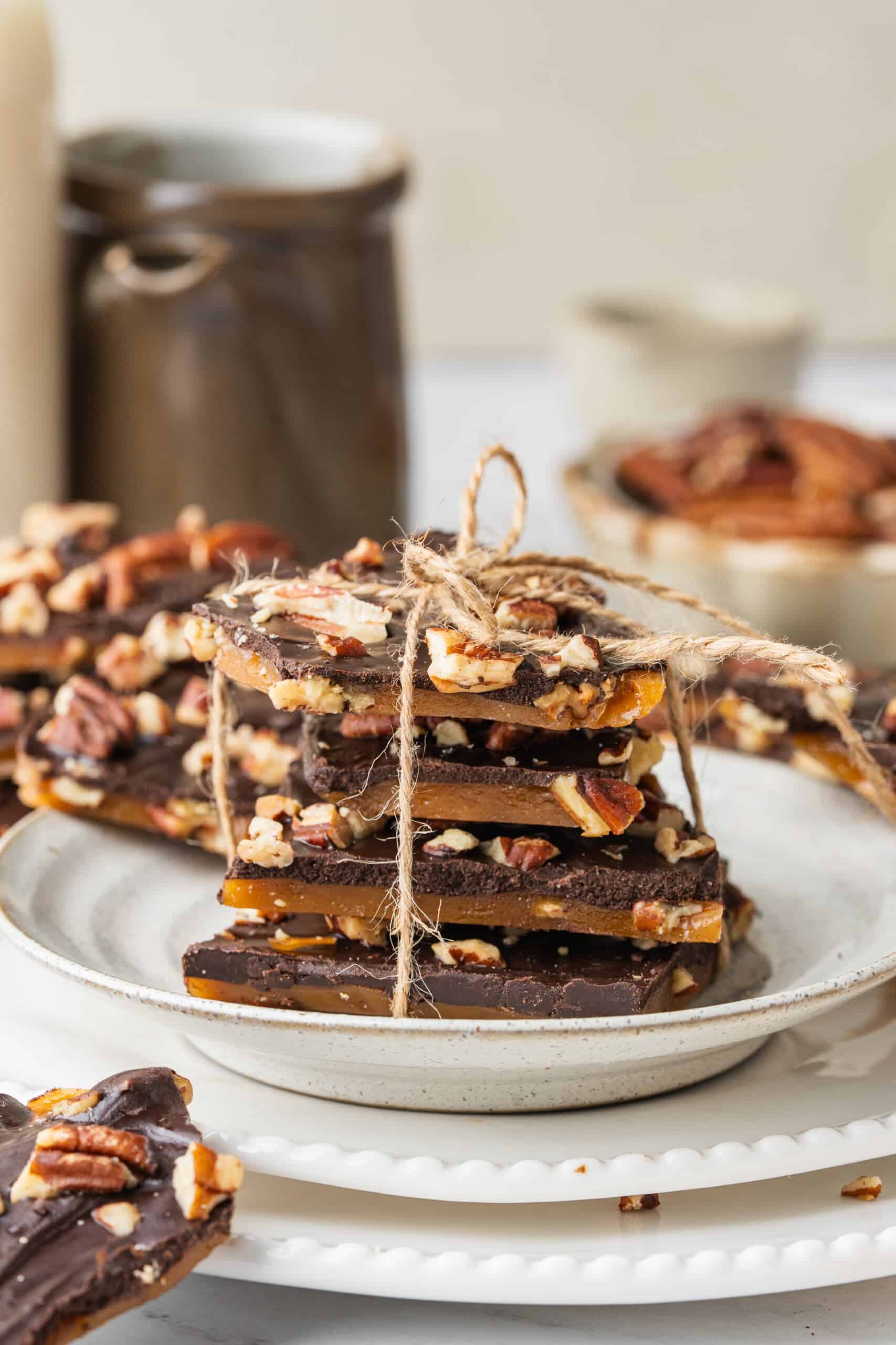 Homemade Toffee with layers of buttery caramel, dark chocolate, and chopped pecans stacked and tied with rustic twine on a ceramic plate.