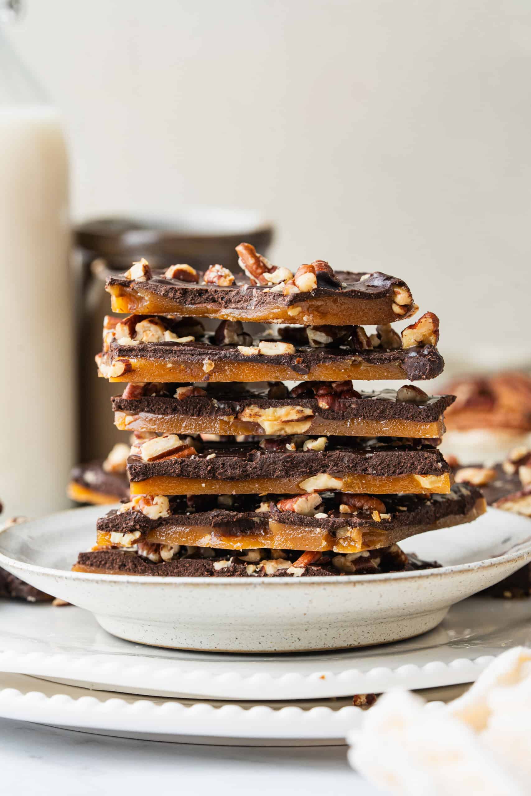 Homemade Toffee stacked on a ceramic plate, showing layers of golden buttery toffee, dark chocolate, and chopped pecans.