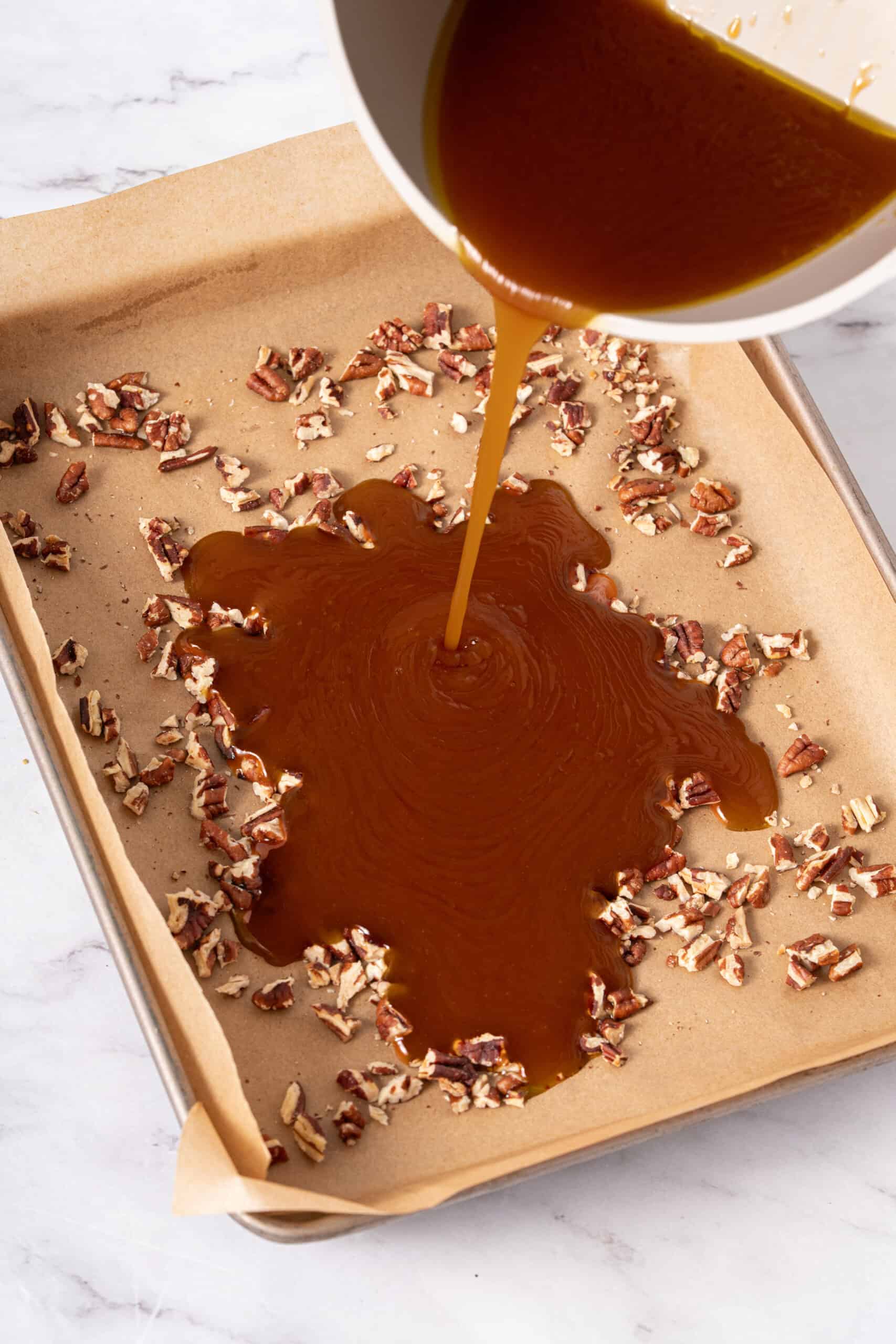Hot golden-brown sugar mixture being poured over chopped pecans on a parchment-lined baking sheet.