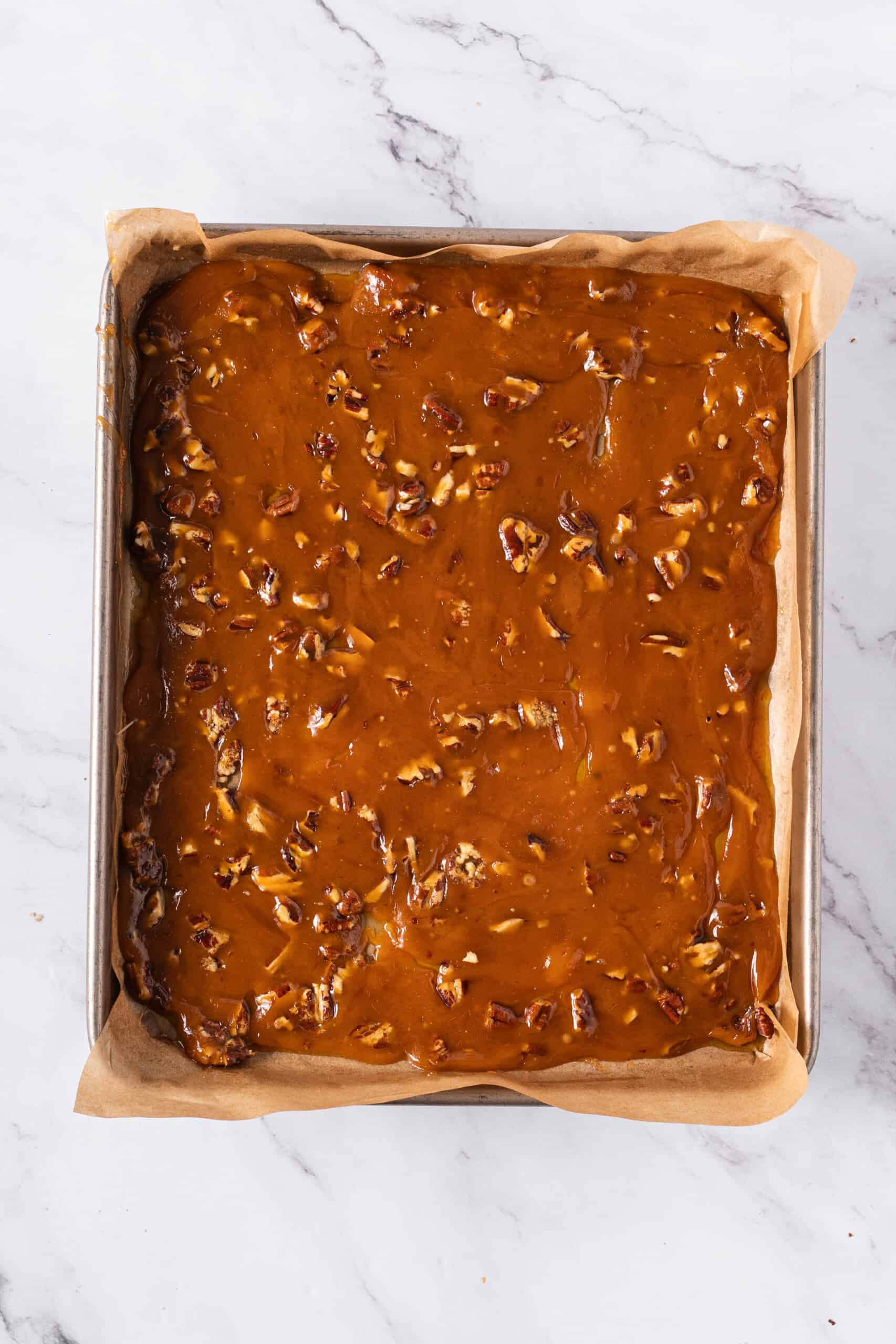 Golden-brown sugar mixture spread evenly over chopped pecans on a parchment-lined baking sheet.