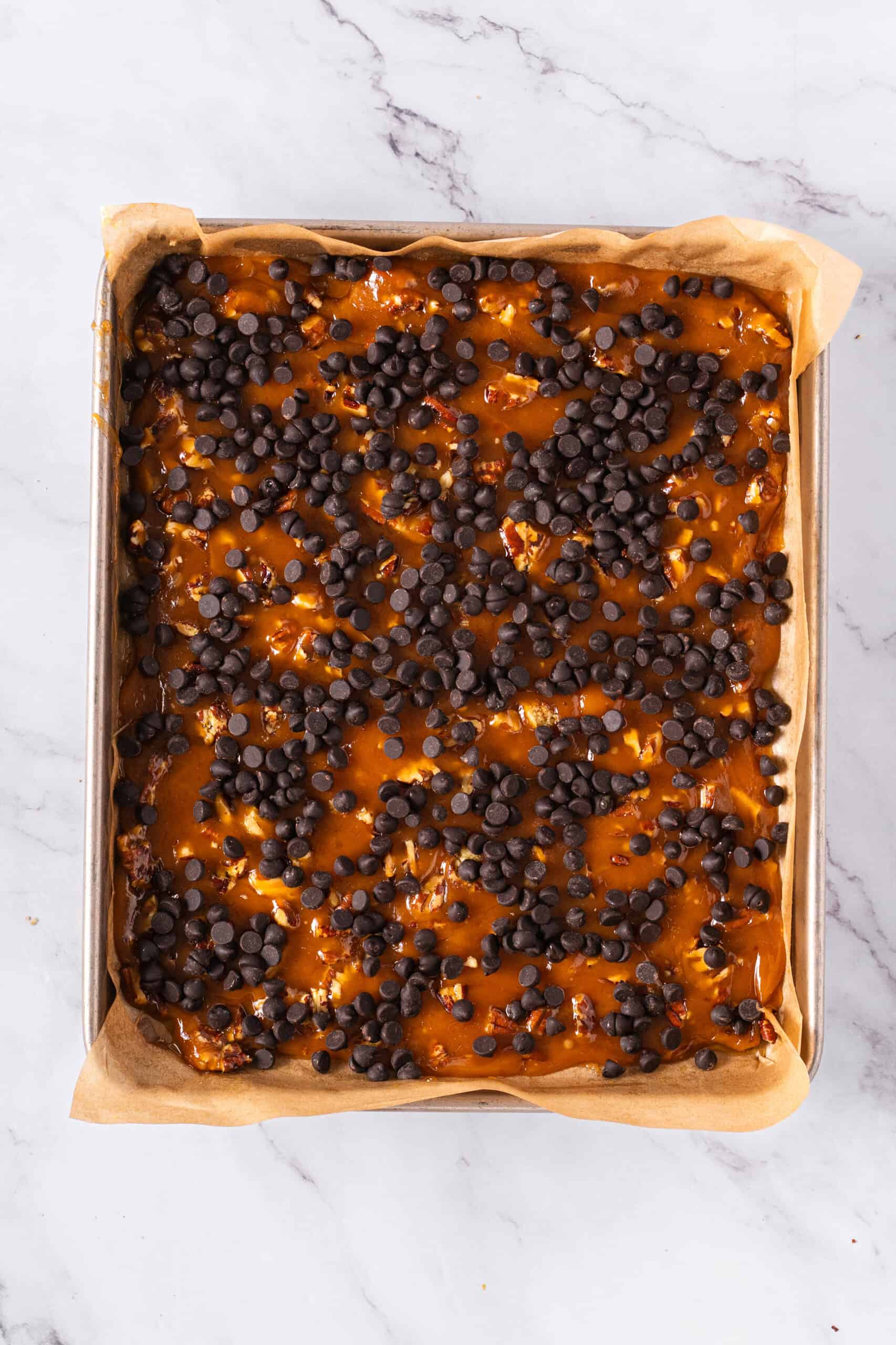Chocolate chips sprinkled over a golden-brown butter and sugar mixture with chopped pecans on a parchment-lined baking sheet.