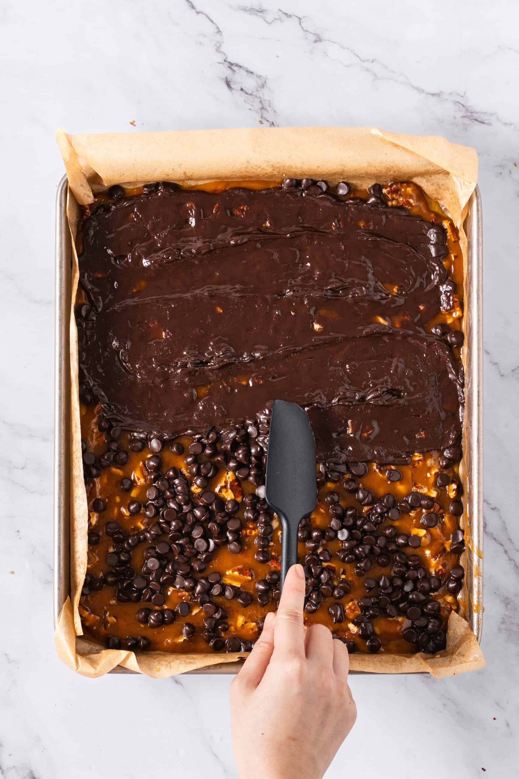Using a spatula to spread melted chocolate over the golden-brown sugar mixture on a parchment-lined baking sheet.