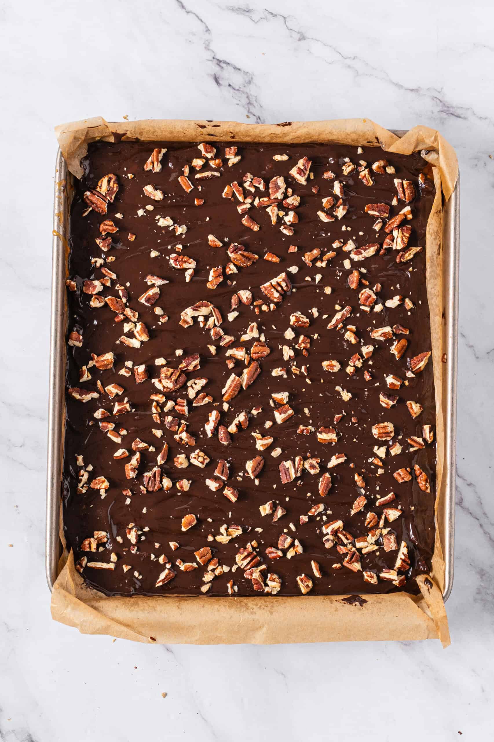 Chopped pecans sprinkled over a smooth layer of melted chocolate on a parchment-lined baking sheet.